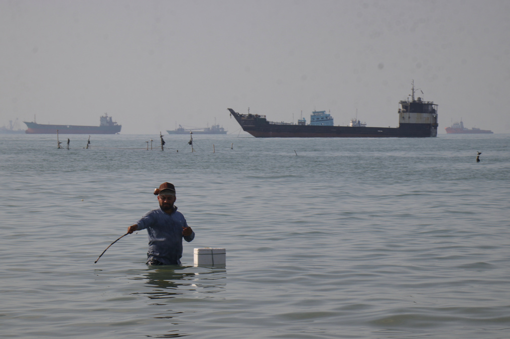 A man stands in the water, appearing to fish, as bulk carriers, cargo ships, and service vessels line the horizon in the Strait of Hormuz off Bandar Abbas, Iran, Monday, April 27, 2026.