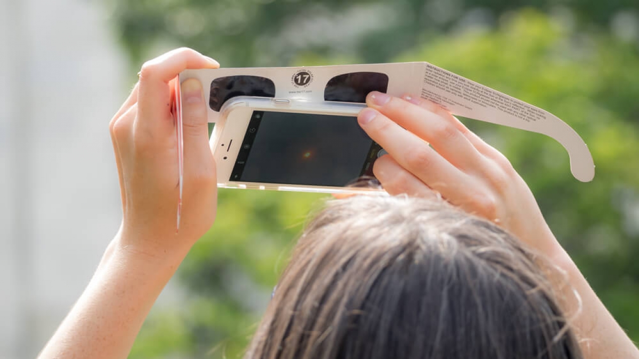 A woman taking a picture of a partial solar eclipse using her smartphone and a pair of solar eclipse glasses.