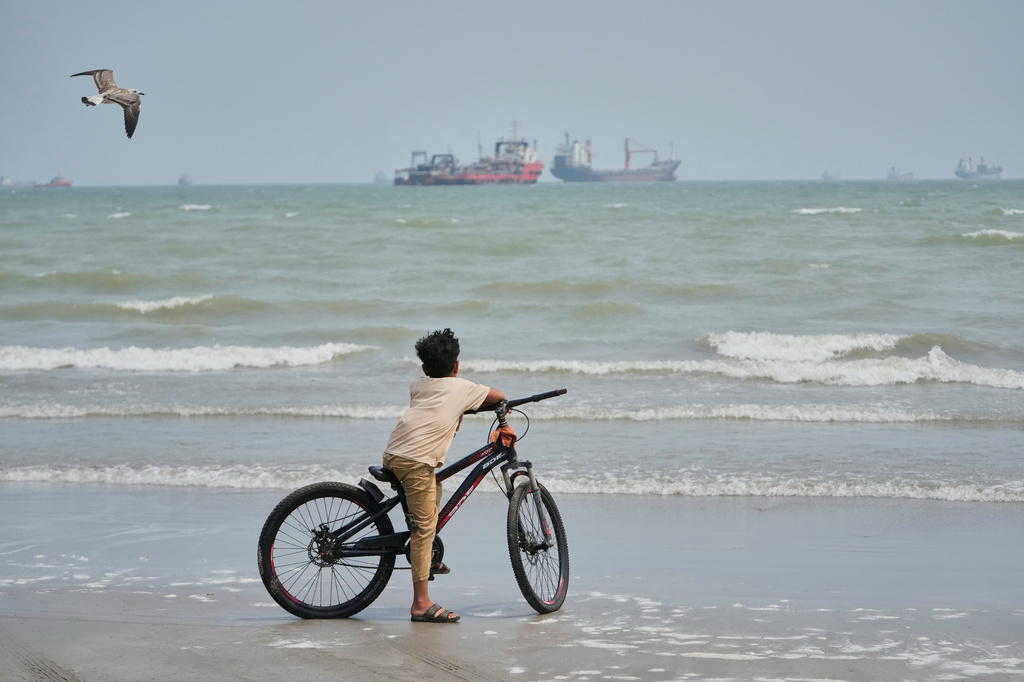 A gull flies overhead as a boy stands with his bicycle on a beach overlooking commercial vessels anchored in the Strait of Hormuz off Bandar Abbas, Iran, Monday, Sept. 7, 2026.