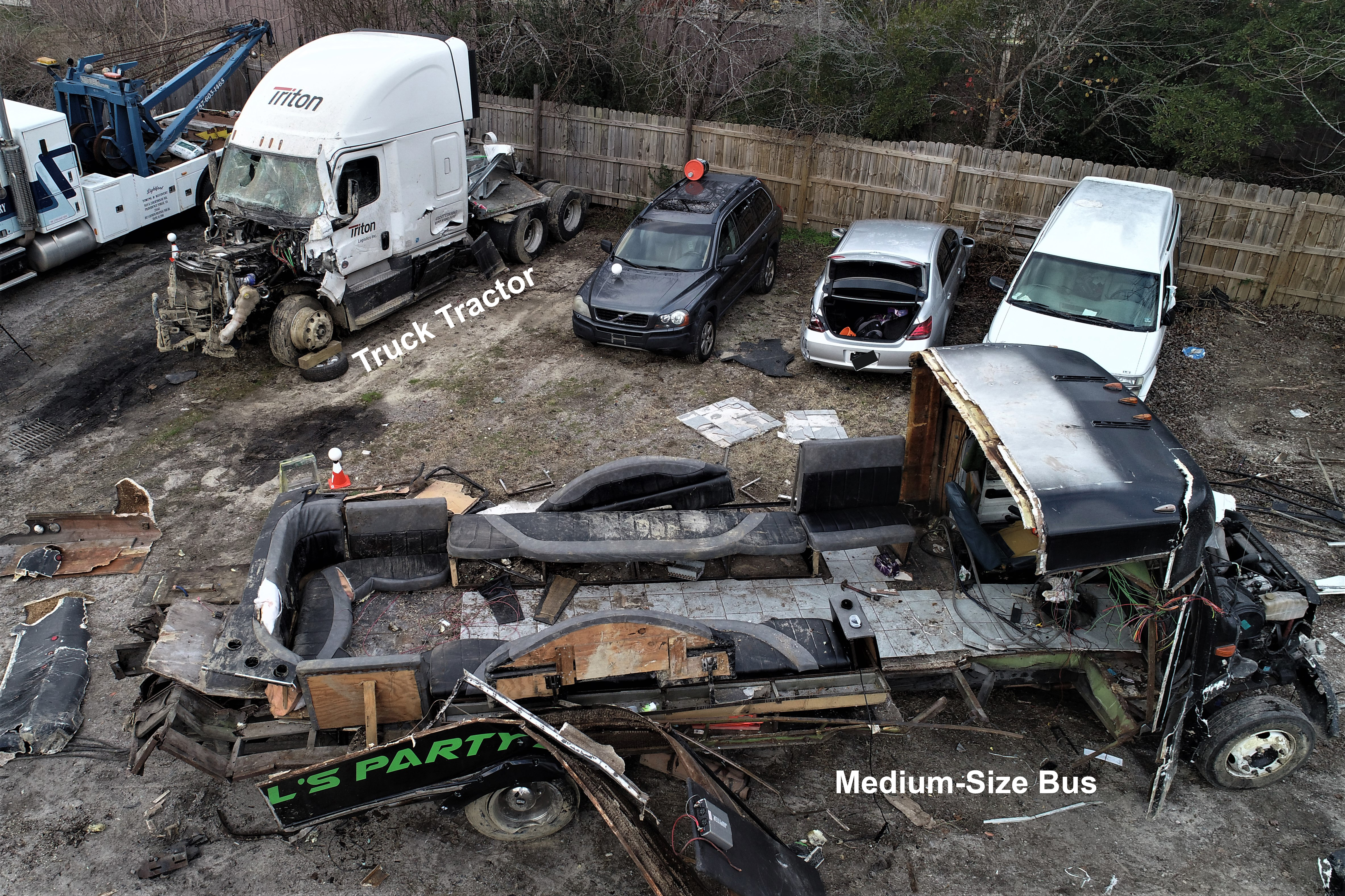 An elevated view showing both vehicles after they had been removed from the crash scene and taken to a tow yard. The medium-size bus is in the foreground. The bus body sustained catastrophic damage in the crash and the interior seating can be seen from this right-side view of the vehicle. The truck-tractor from the combination vehicle is located behind and to the left of the bus, and the significant damage to the front of the vehicle can be seen.