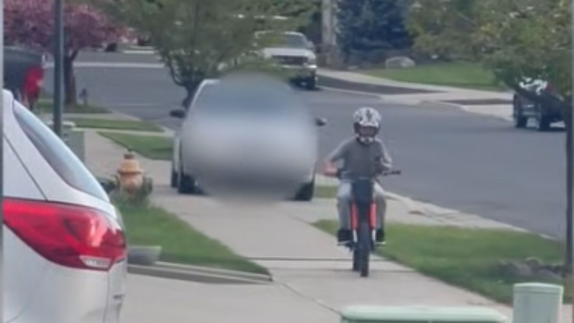A car is seen driving on the sidewalk behind a child who is riding an electric bike.