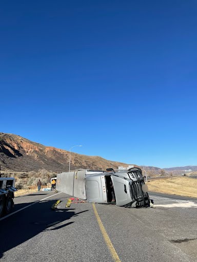 The semi rolled over on to its side, the cabin of the vehicle partially dented under its own weight. It is morning. Two people are seen in the background assessing the damage.