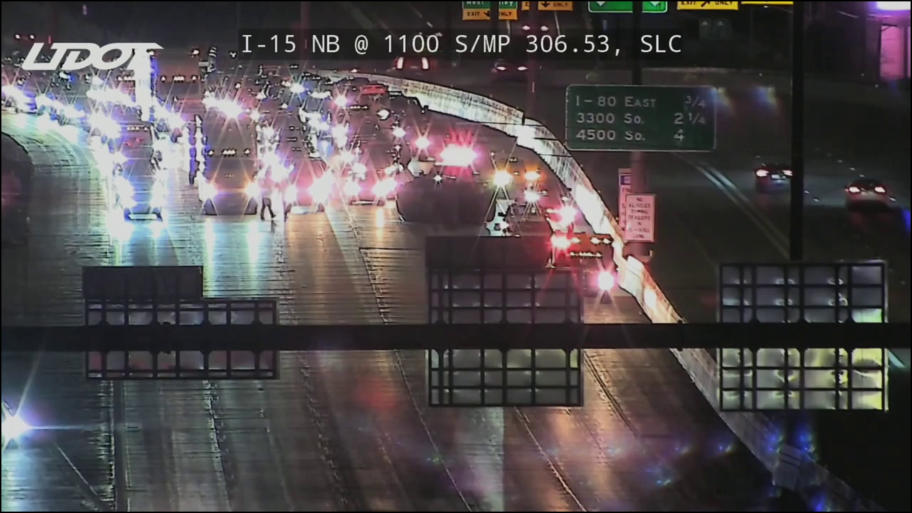 A still from a UDOT Traffic Camera of the scene. It's dark, and headlights flood the freeway with light. Flashing emergency vehicles block off traffic as a trooper walks nearby.