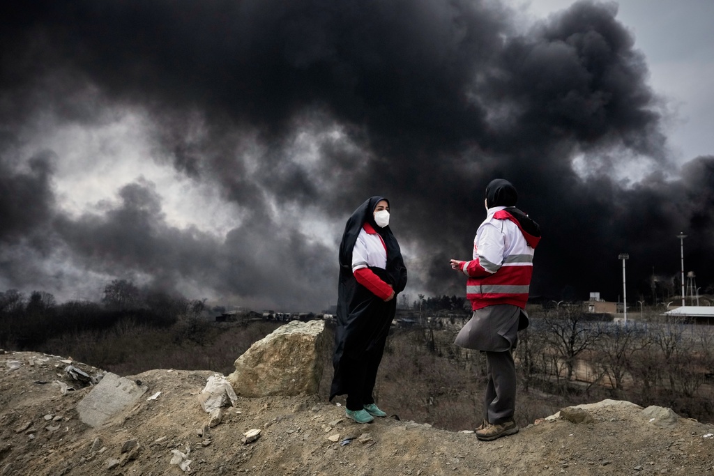 Two women from the Iranian Red Crescent Society stand as a thick plume of smoke from a U.S.-Israeli strike on an oil storage facility late Saturday rises in the sky in Tehran, Iran.