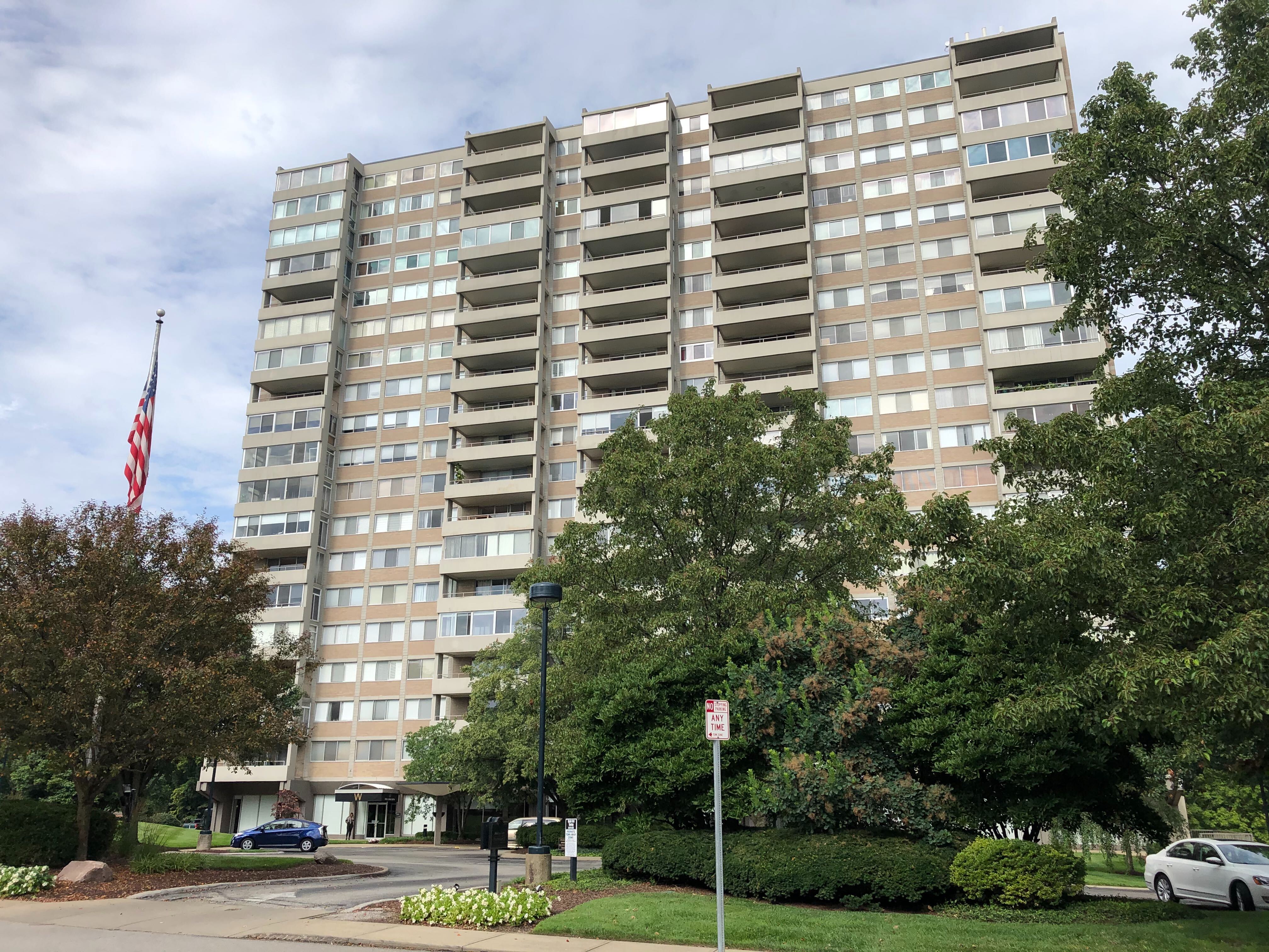 A 19-story apartment tower, beige in color, checkered with windows and balconies, sits behind a line of trees and in front of a partly cloudy sky.