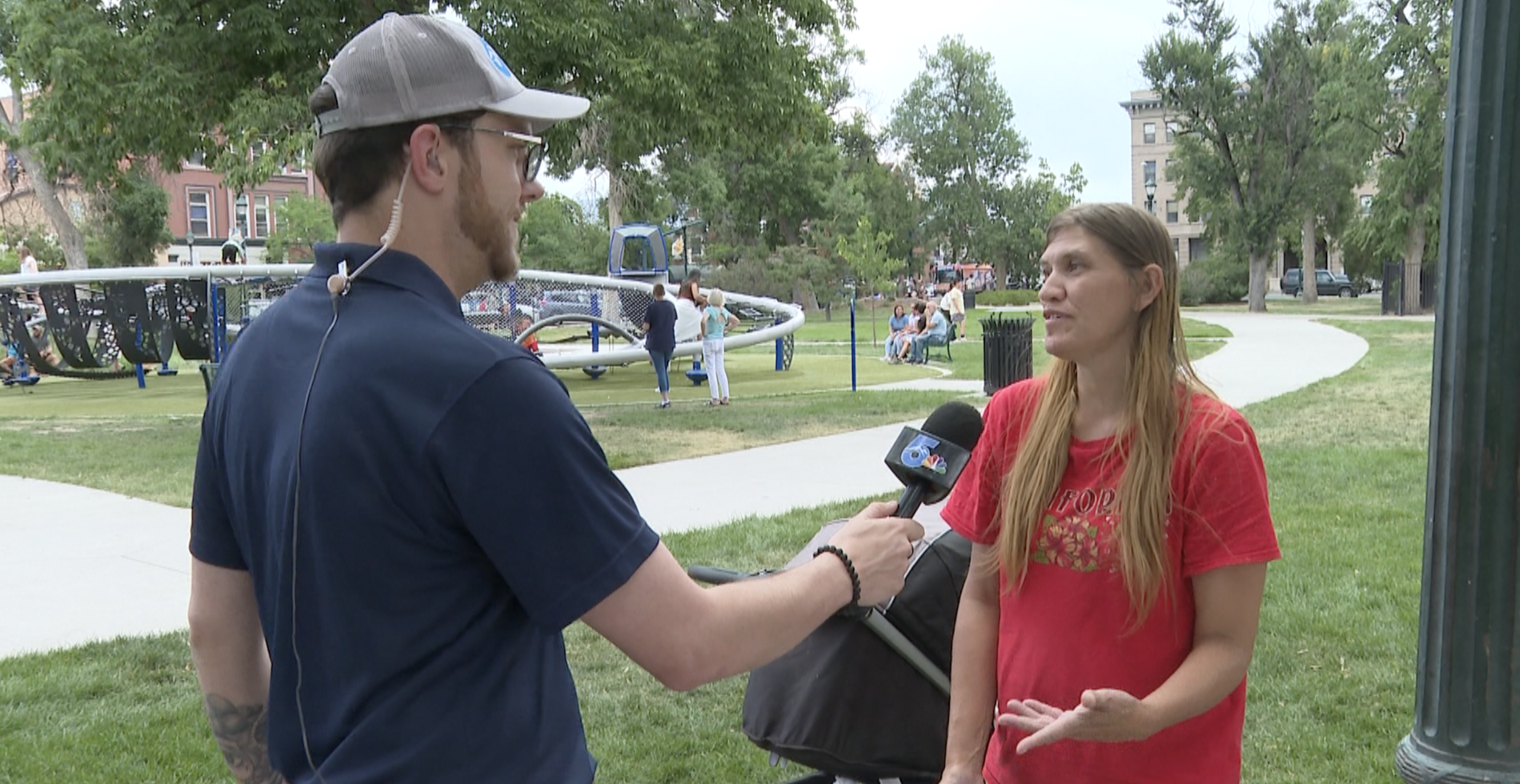 A photo of reporter Jake Walker interviewing a woman at a park. He's holding a News 5 Microphone and wearing blue. She's wearing red and has blonde hair. You can see playground equipment in the background.