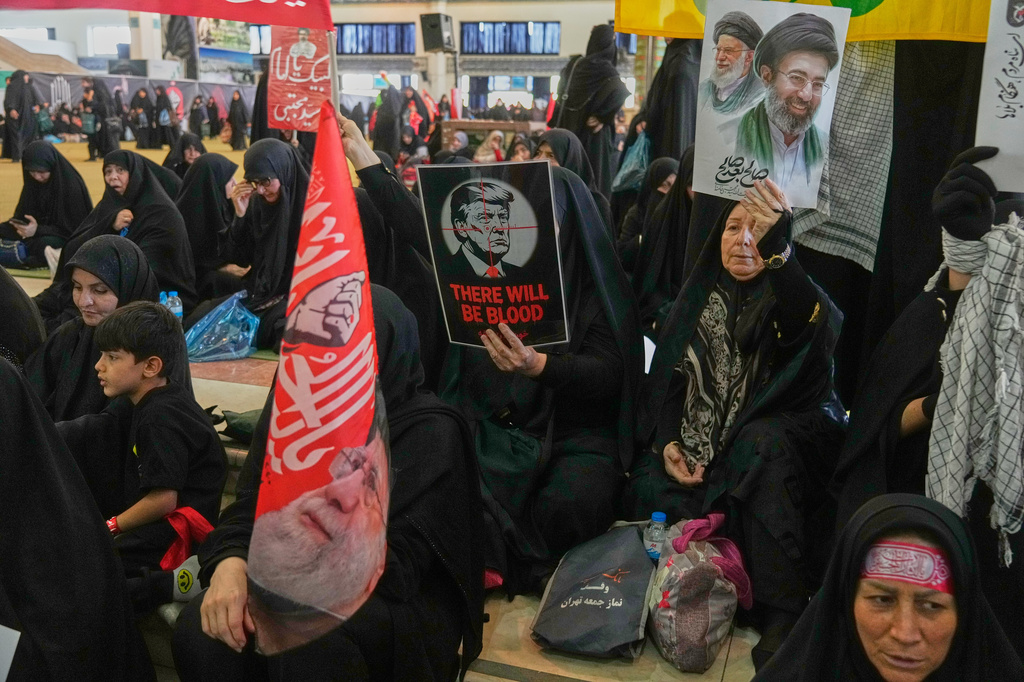 A woman holds a poster depicting U.S. President Donald Trump in the crosshairs of a rifle scope with the English words "There Will Be Blood," while another woman holds a portrait of the late Supreme Leader Ali Khamenei and his son, current Supreme Leader Mojtaba Khamenei, as mourners gather to commemorate the late leader at the Imam Khomeini Mosalla Grand Mosque in Tehran, Iran, Tuesday, July 14, 2026. 