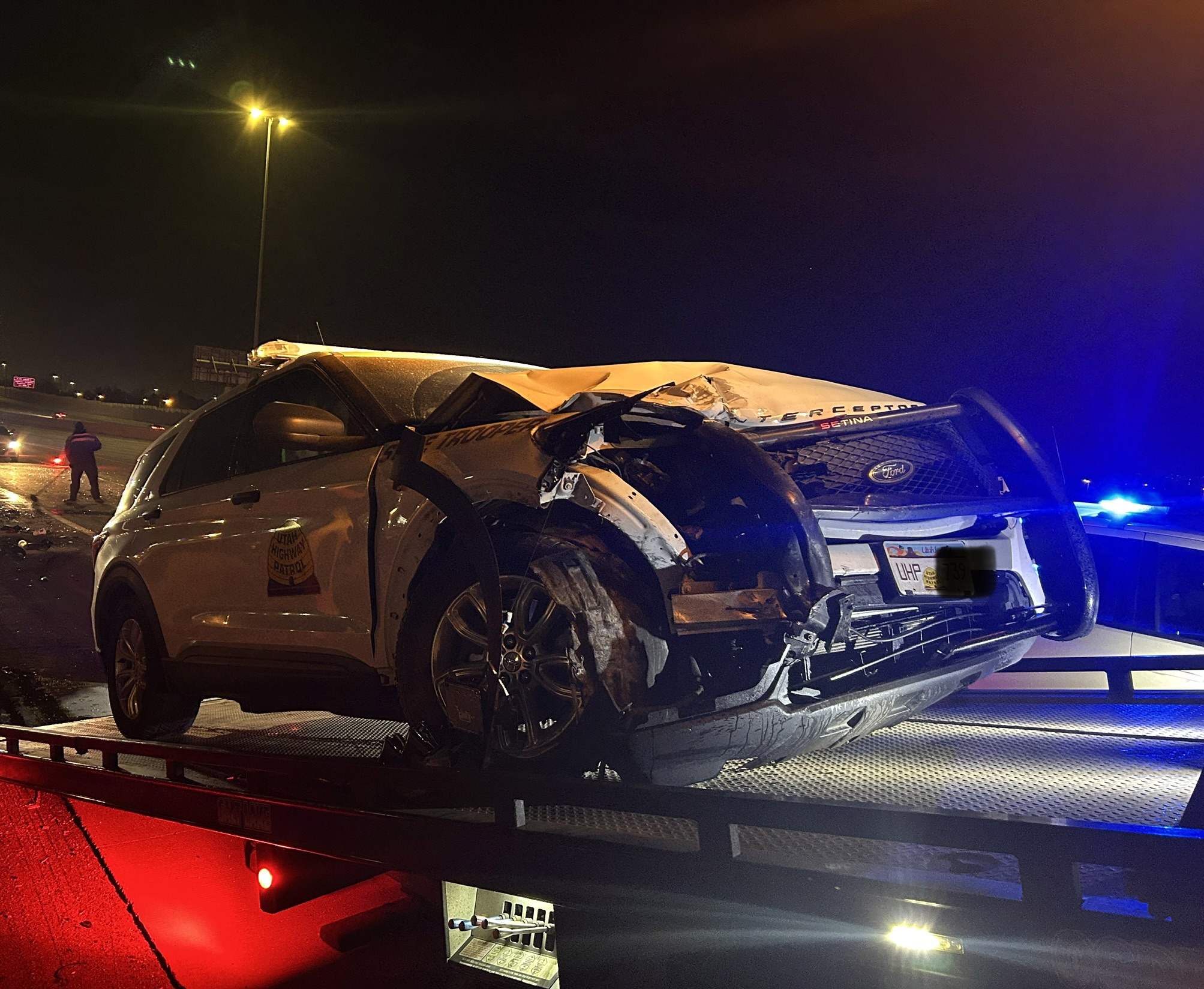 A photo of the scene released by Utah Highway Patrol. It's night. The damaged patrol vehicle is hoisted onto the bed of a tow truck. It shows visible front end damage.