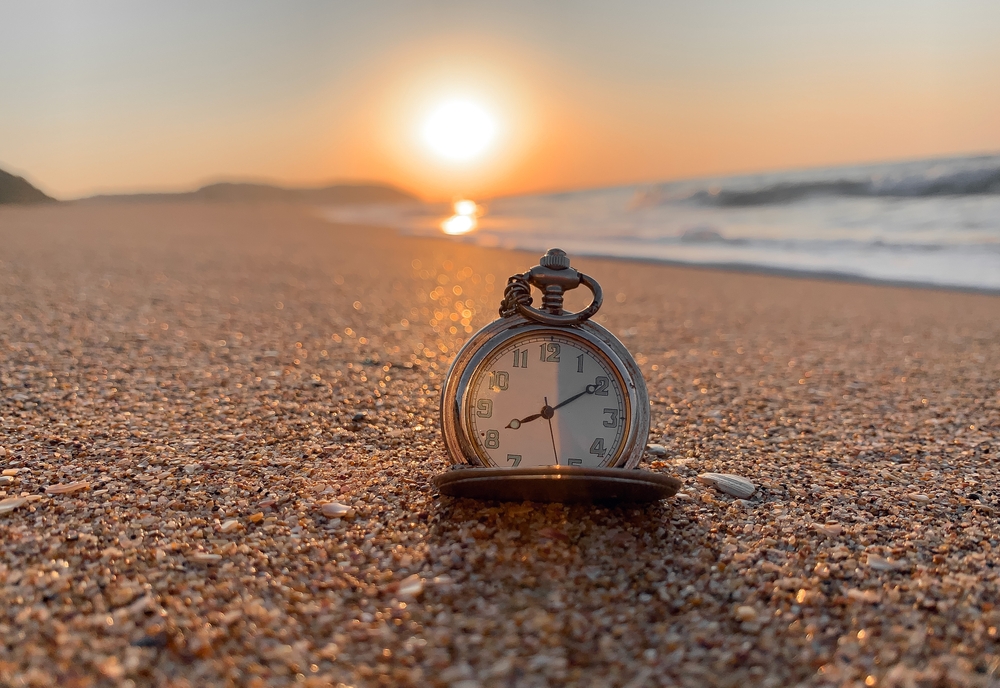 A clock sits on a beach in front of a setting sun.