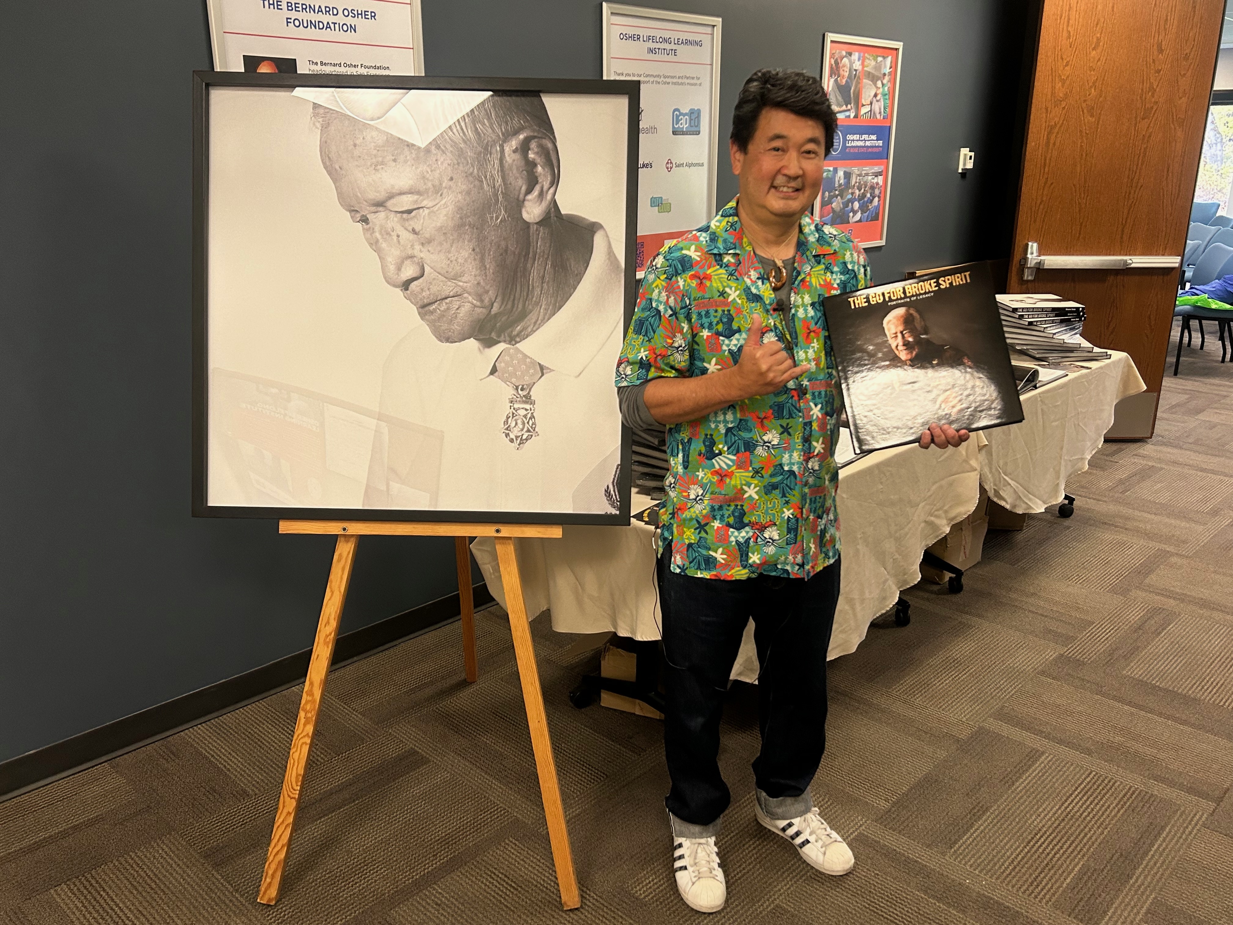 Shane Sato stands beside one of his photographs while holding his book 'The Go for Broke Spirit.'