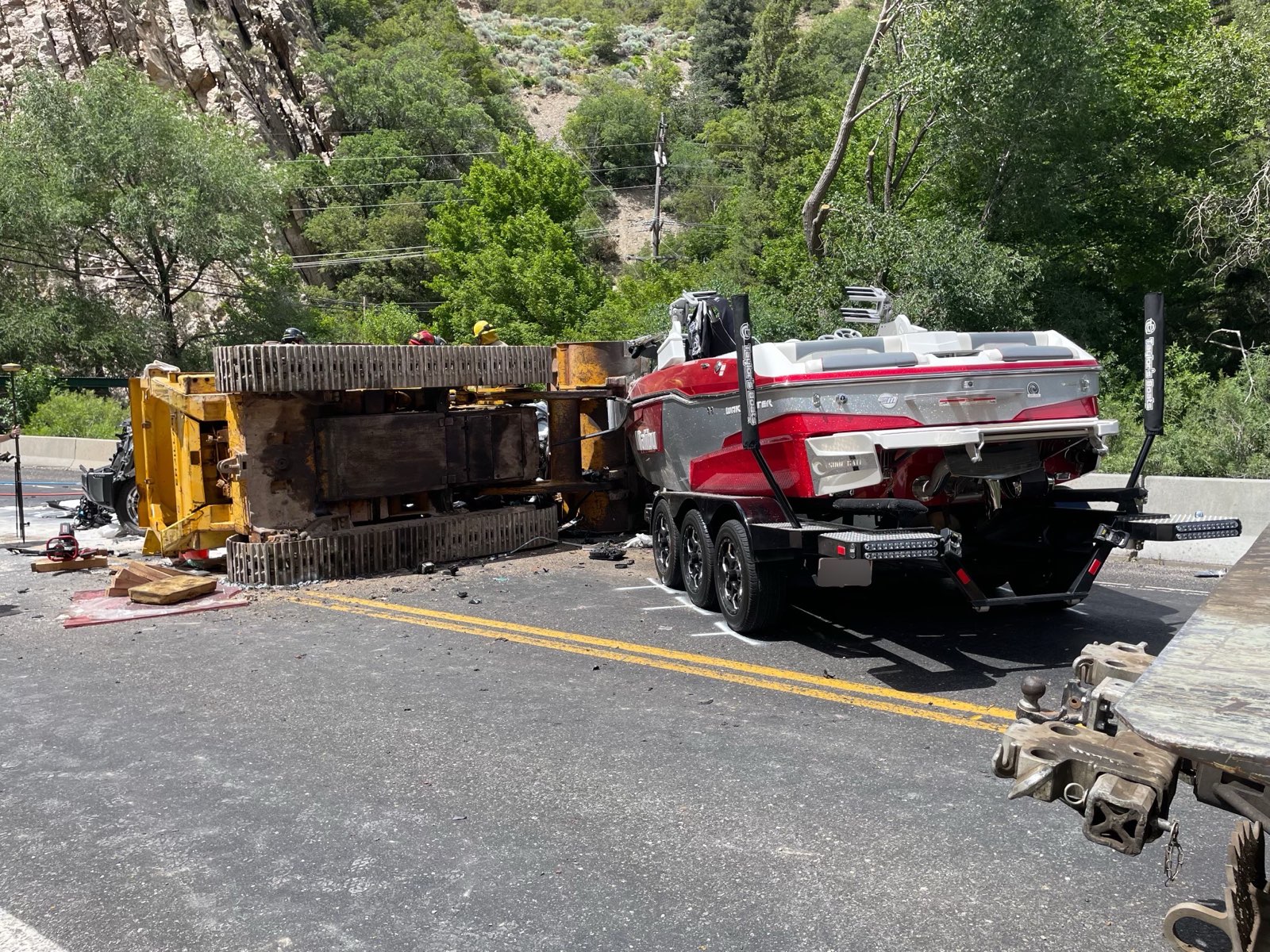 A photo of the scene released by Utah Highway Patrol. It's daytime. On the left side of the image, what appears to be a bulldozer is lying on its side. Next to it is a trailer that was hauling what appears to be a red speedboat.
