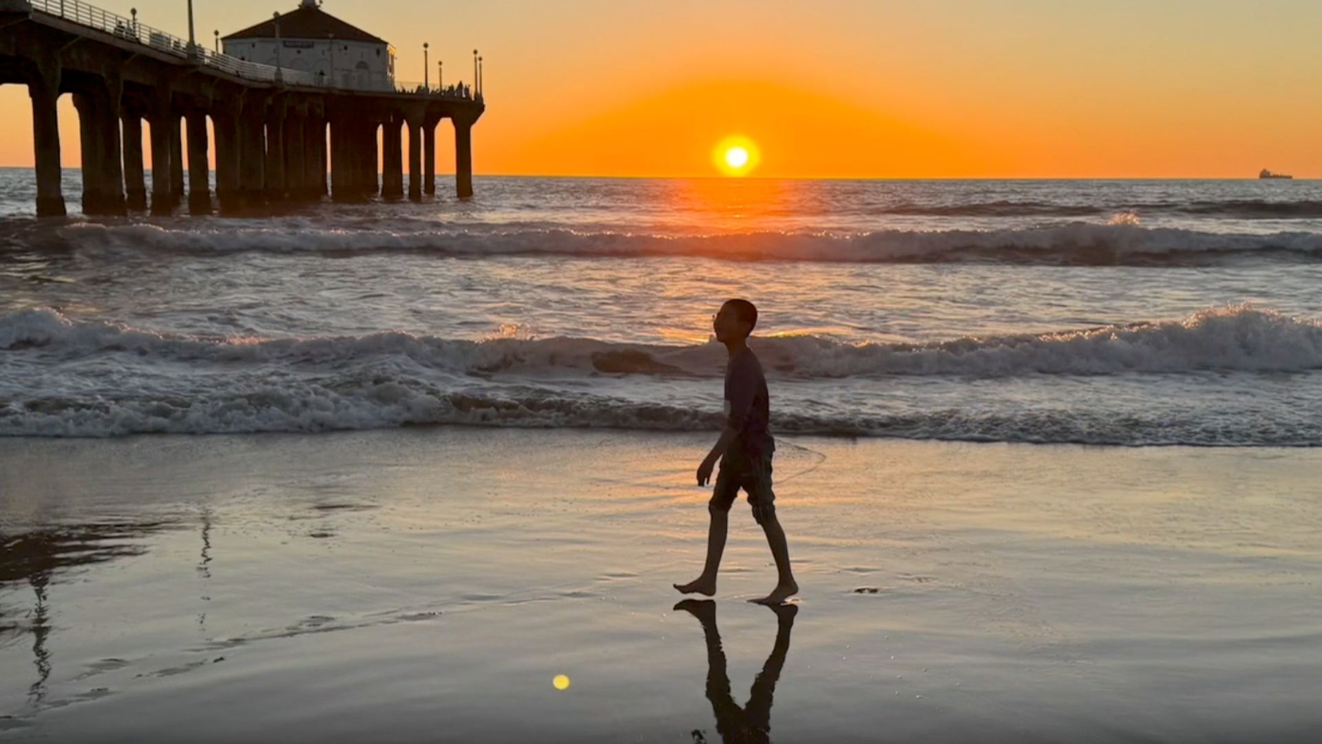 A person walks along Manhattan Beach in California as the sun sets over the water in the background.