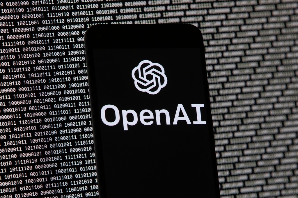 The OpenAI logo appears on a mobile phone in front of a computer screen with random binary data, March 9, 2023, in Boston. 