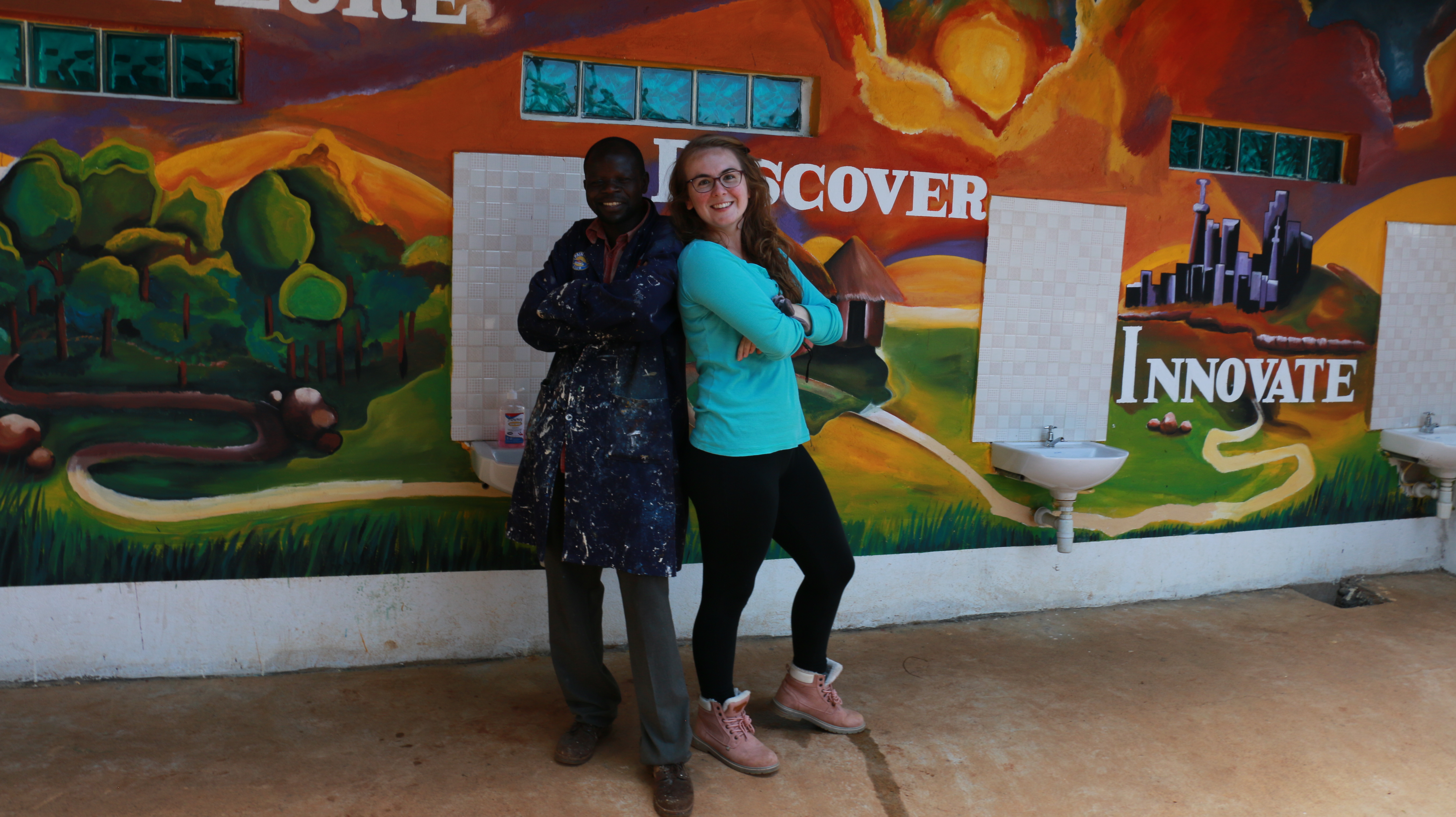 A man and a woman pose with their arms crossed and backs touching in front of a mural of the Western Kenyan landscape 