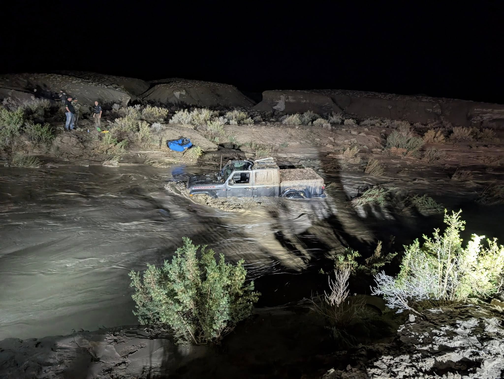 An image from the rescue released by the Grand County Sheriff's Office. It's nighttime. The scene is light with lights from emergency vehicles. A black pickup truck sits in the middle of the creek, covered in mud. The water has risen to the level where the tires are fully submerged. A group stands on the far bank in the background, talking among one another.