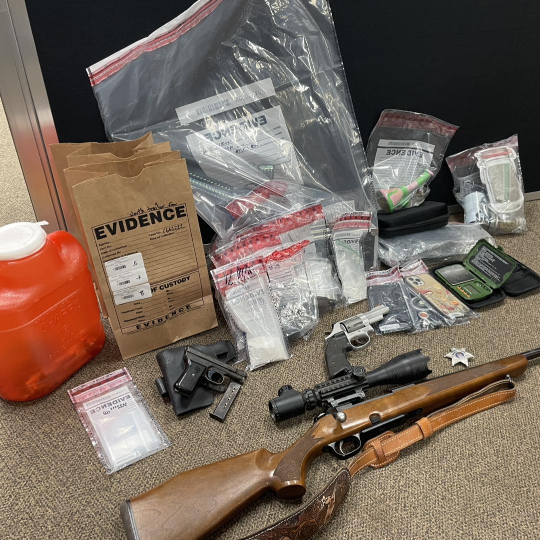 A photo of drugs and firearms seized by the Grand County Sheriff's Office. Many of the smaller items are in bags labeled "EVIDENCE". At the front of the drugs and paraphernalia are three guns, a rifle with a scope, a hand gun, and a revolver.