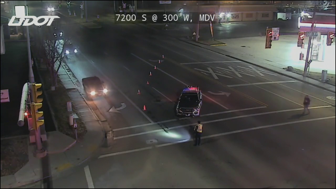 A still taken from a UDOT Traffic Camera. It's dark. Traffic is reduced to one lane, and a police officer in a reflective vest uses a flashlight to direct traffic.