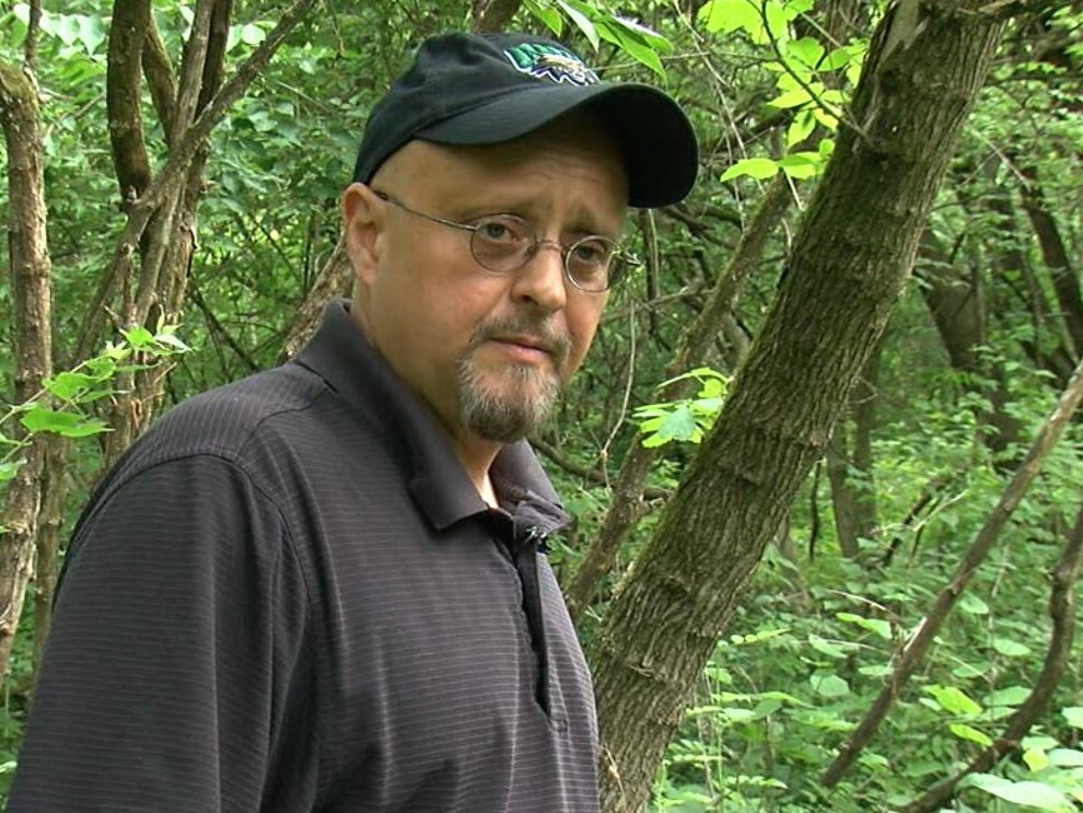 Michael Morgan is pictured here standing in the overgrown Potter's Field cemetery. He is wearing a black Ohio University baseball cap and a black shirt.