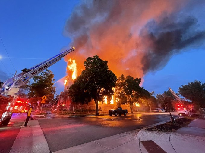 A photo of the scene released by Salt Lake City Police. It's early morning, the sun has begun to rise. The chapel is fully engulfed. Smoke billows into the sky as multiple fire engines work to battle the fire.