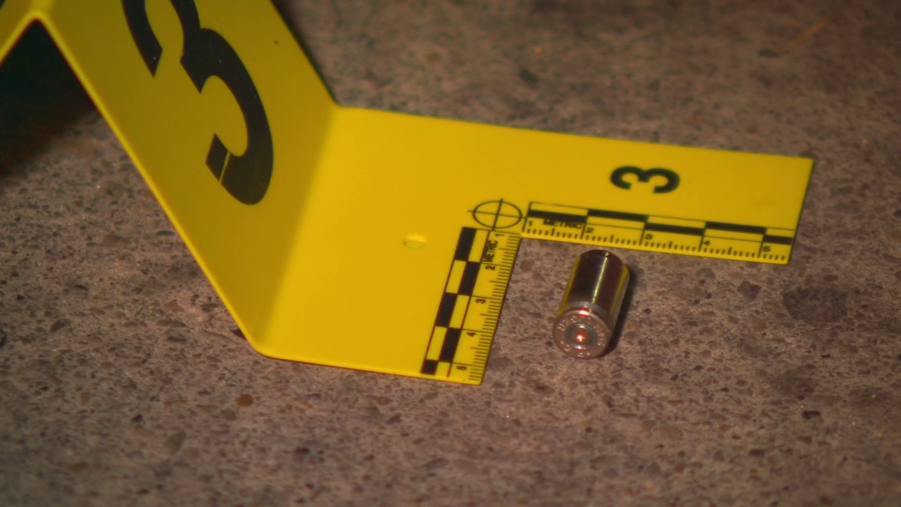 An image of the scene taken by a FOX 13 photojournalist. It is night, and a crime scene marker is placed next to a bullet casing, which sits on the sidewalk. The marker includes a ruler that indicates the size of the casing, which is about one centimeter wide and about two or three centimeters long.