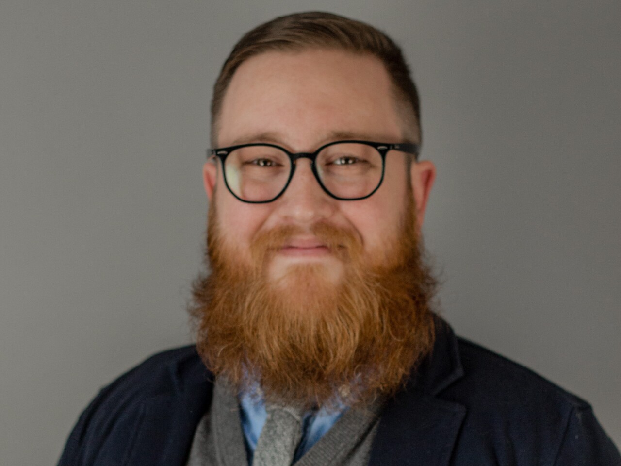 Justin Tucker is smiling in this portrait. He has a full, red beard and mustache and short-cropped hair. He's wearing glasses, a blue shirt and a gray tie with a gray sweater and dark blazer.