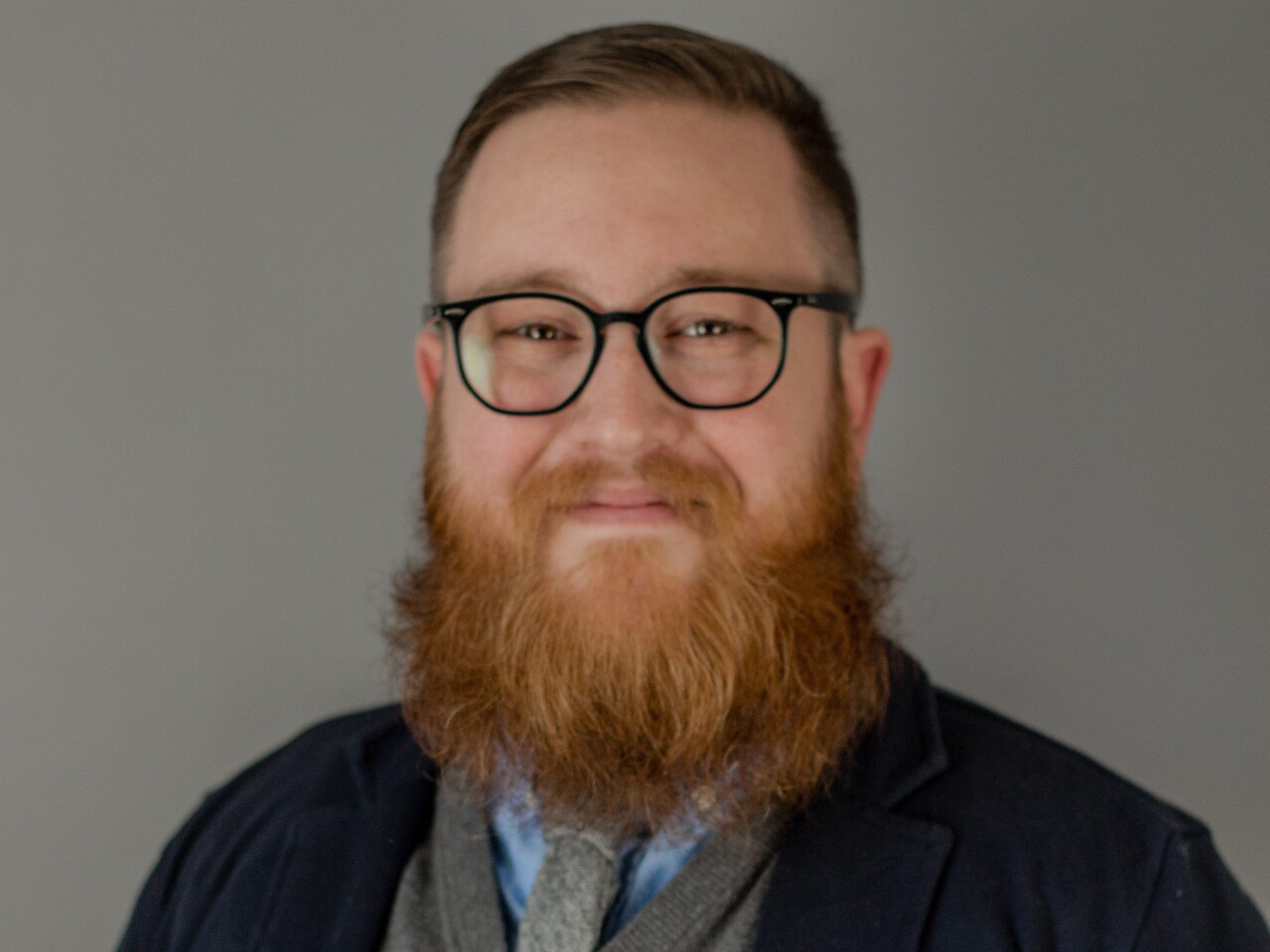 Justin Tucker is smiling in this portrait. He has a full, red beard and mustache and short-cropped hair. He's wearing glasses, a blue shirt and a gray tie with a gray sweater and dark blazer. 