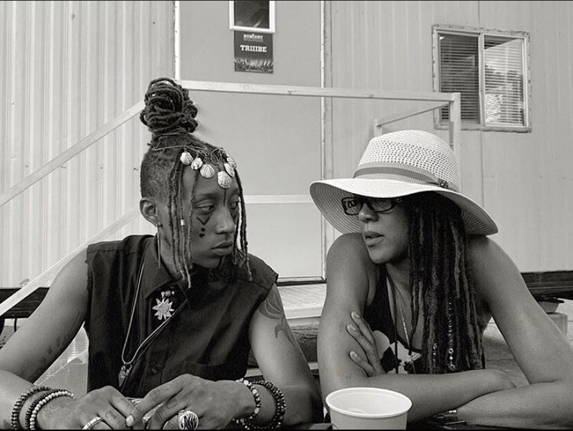 In this undated photo, Siri Imani, on the left, is looking at her mom, Jennie Wright, as they sit next to each other. Imani is wearing her braided hair in a pile on top of her head, and her face is painted. Wright's long braids hang down below her straw hat.