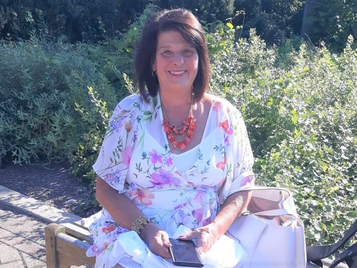 Barbara Fleckenstein smiles in this undated photo. She is seated on a bench. She has dark, shoulder-length hair and is wearing a white, floral print dress.