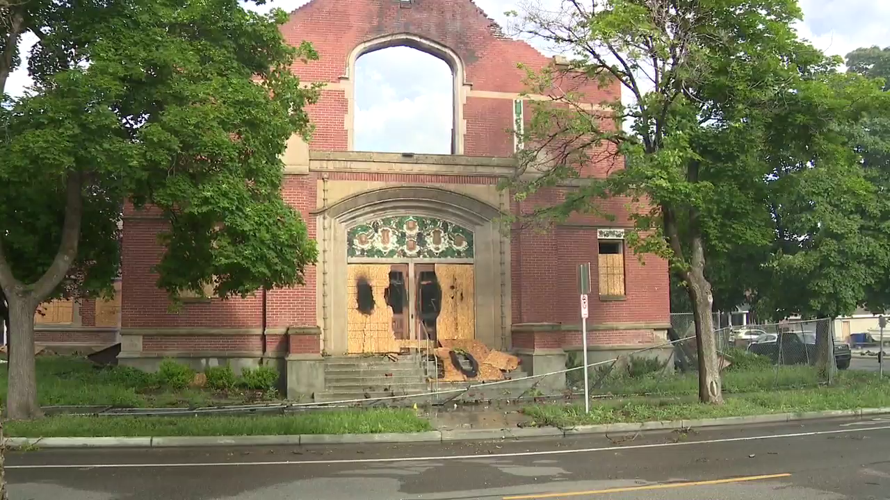A photo of the building taken by a FOX 13 Photojournalist. It's daytime, and we see what used to be the main entrance to the chapel. The wood that boarded up the doors is burned through. The space where a stained glass window once sat lies empty. Bits of burnt debris are scattered on the concrete steps leading to the entrance.