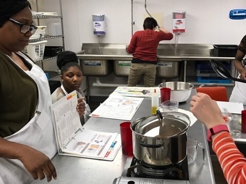 LaTasha Hambrick, left, and her daughter, seated, listen to instructions during a 2018 Cooking for the Family class. Hambrick is wearing glasses and a white apron over a black top.