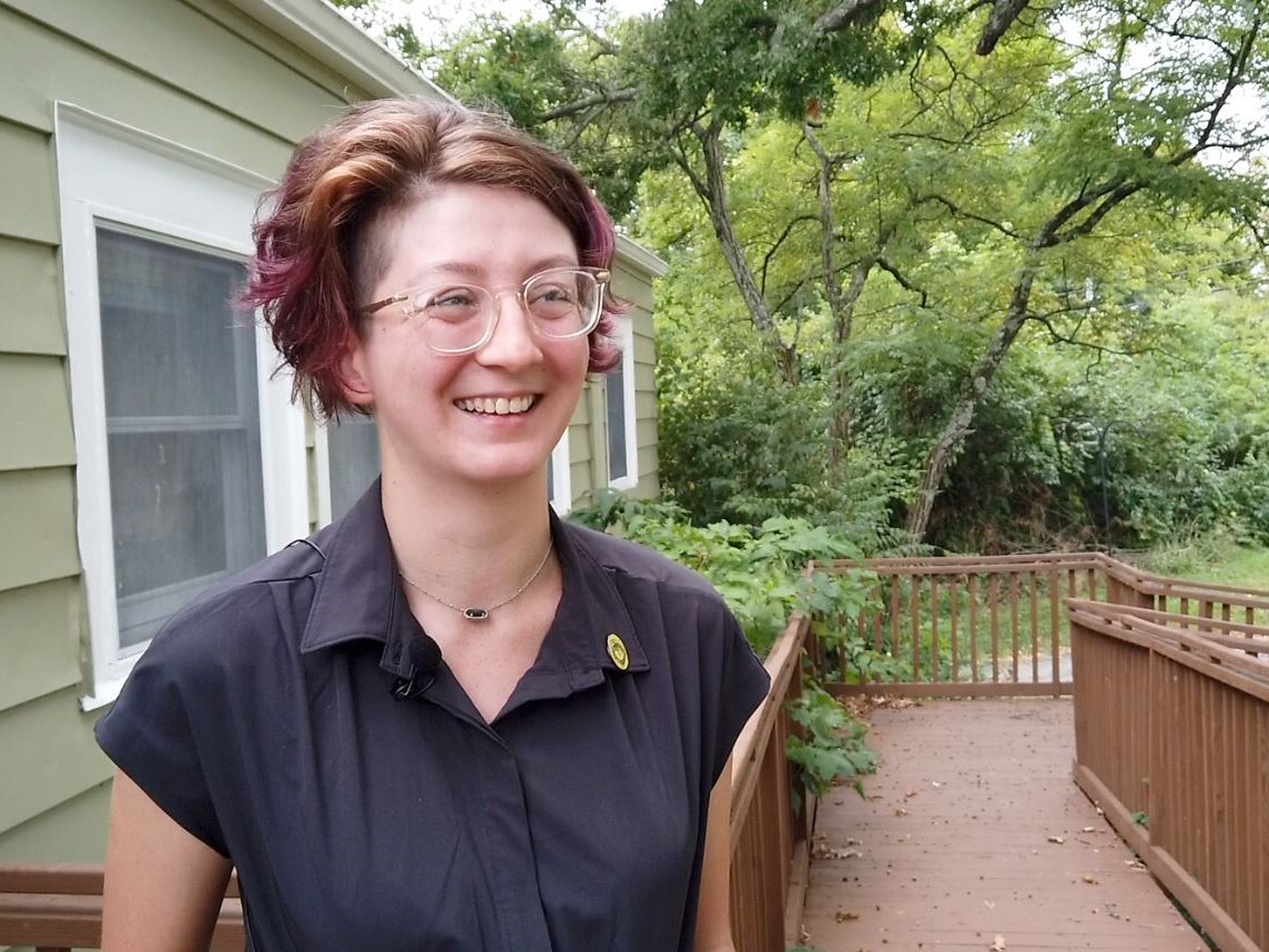 Bethany Heeg stands outside Shine Nurture Center in Mt. Airy. She is smiling and wearing glasses, and her wavy brown hair has some purple color in it.