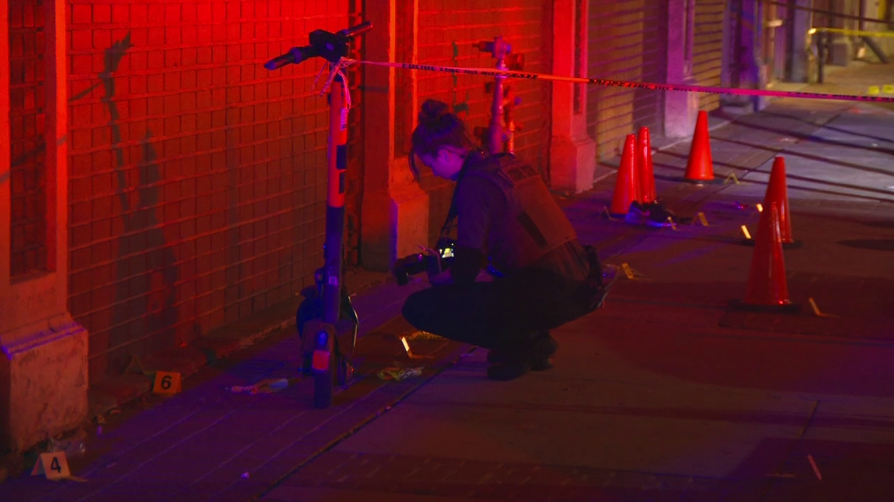 An image of the scene shot by a FOX 13 Photojournalist. It is night. An investigator with Salt Lake City Police's forensic lab takes photos and documents evidence of the shooting found at the scene. She is surrounded by a number of cones and markers that map out the scene, with crime scene tape anchored on an electric scooter. The scene is lit by the flashing red and blue lights of a nearby patrol vehicle.
