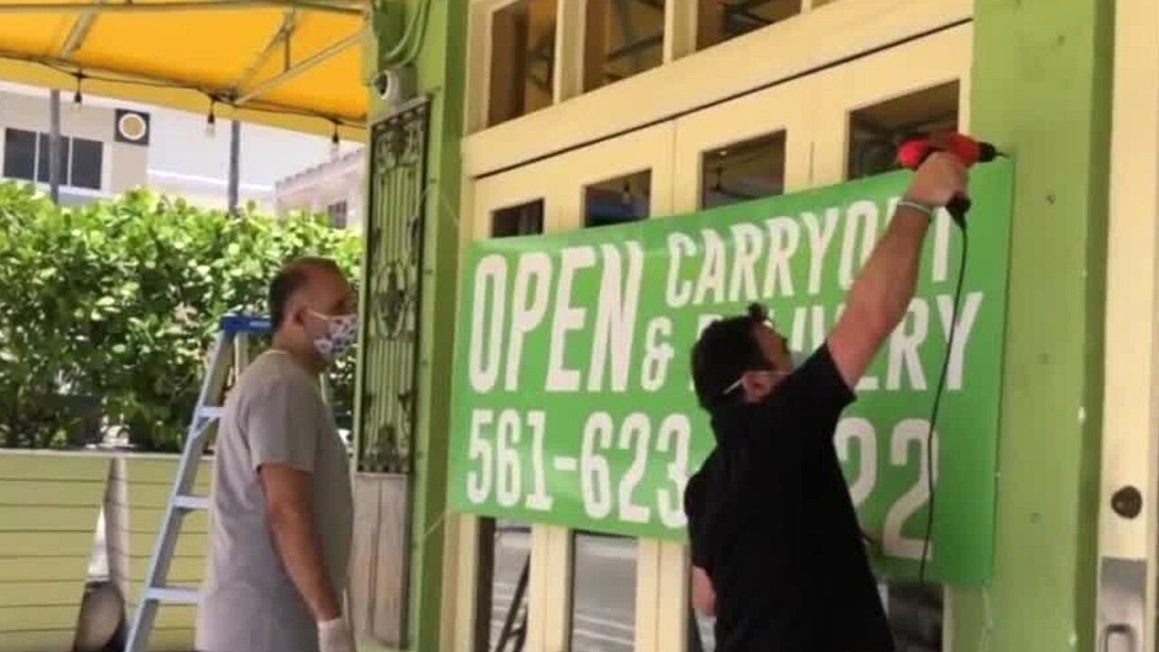 Workers hang a sign outside of Avocado Grill in West Palm Beach on April 23, 2020 to indicate the business now offers carryout and delivery.