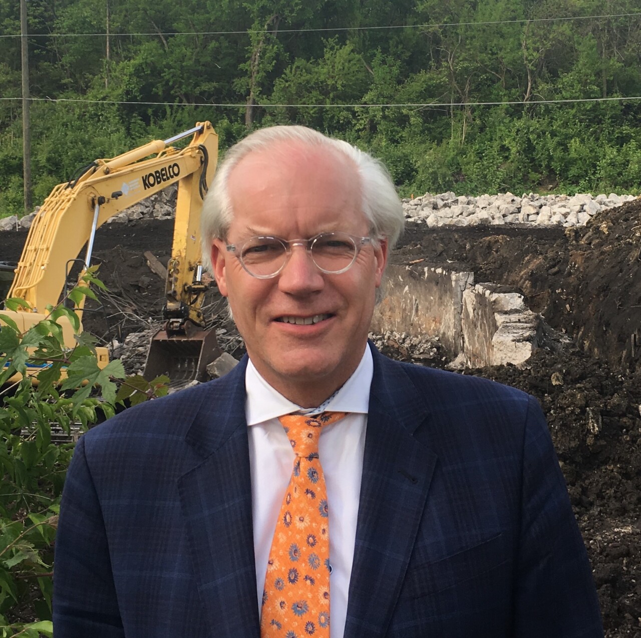 T.J. Ackermann poses for a photo at a construction site. He has white hair and glasses. He's wearing a white shirt, a yellow patterned tie and a navy, patterned jacket.