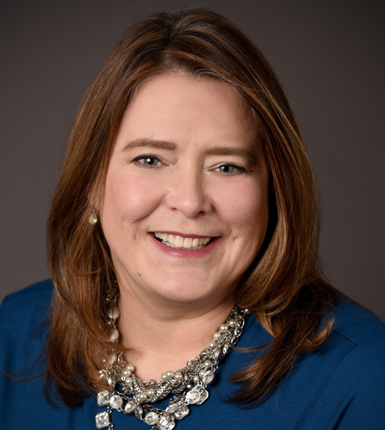 Shannon Starkey-Taylor smiles in this portrait. She has long, straight brown hair and is wearing a blue top and silver, jeweled necklace.