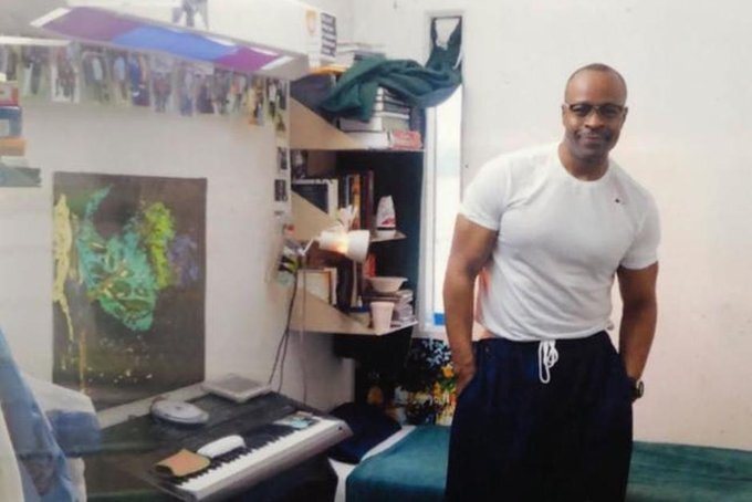 Keith LaMar stands in his prison cell in this undated photo. He has short-cropped hair and glasses and is wearing a white t-shirt and dark pants. His cell is decorated with colorful artwork and photos, and his book shelves are packed with books.
