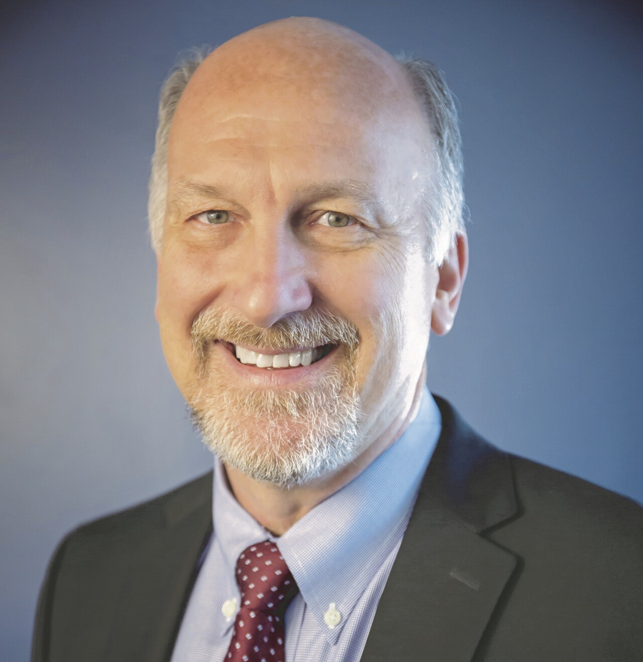 Jock Pitts is smiling in this portrait. He has a grey beard and mustache and grey hair cropped close. He's wearing a tie, button-down shirt and suit jacket.