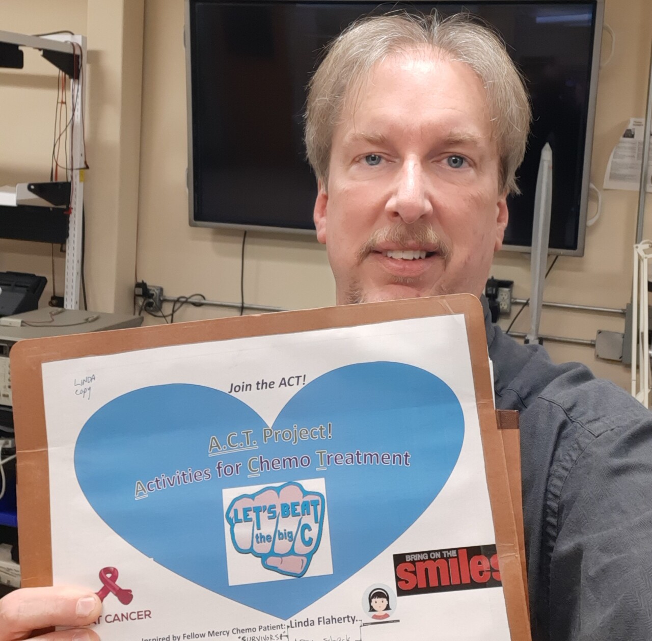 Scott Murphy is smiling in this photo as he holds a folder of activity sheets. He has short grey hair, a beard and mustache. 