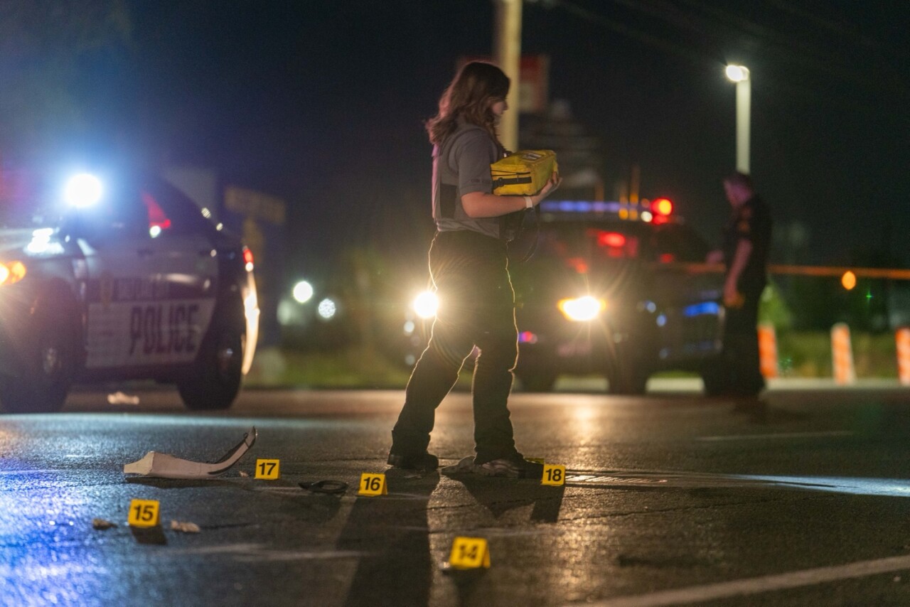 Another scene photo from Salt Lake City Police. It's night. A Salt Lake City Police Crime Lab technician walks around the scene. Evidence markers are on the road. She is carrying what appears to be a bag for collecting evidence in her arms. Two police cruisers are in the background, lights flashing.