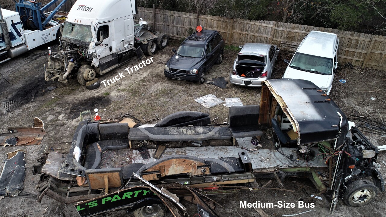 An elevated view showing both vehicles after they had been removed from the crash scene and taken to a tow yard. The medium-size bus is in the foreground. The bus body sustained catastrophic damage in the crash and the interior seating can be seen from this right-side view of the vehicle. The truck-tractor from the combination vehicle is located behind and to the left of the bus, and the significant damage to the front of the vehicle can be seen.