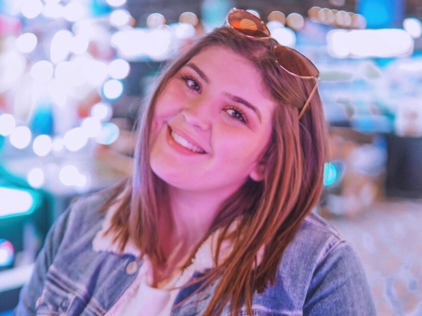 Kaelynn Speed smiles in this photo. She has shoulder-length brown hair, sunglasses on top of her head and is wearing a white top and denim jacket.