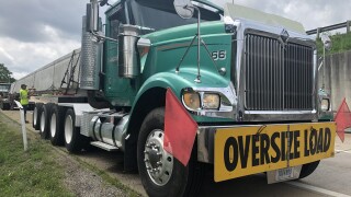 A green semi-trailer truck is parked on the shoulder of an interstate off-ramp. Two red flags are attached to the truck's front fender, along with a yellow sign, reading "Oversize load." A worker wearing a neon yellow shirt stands in the background.