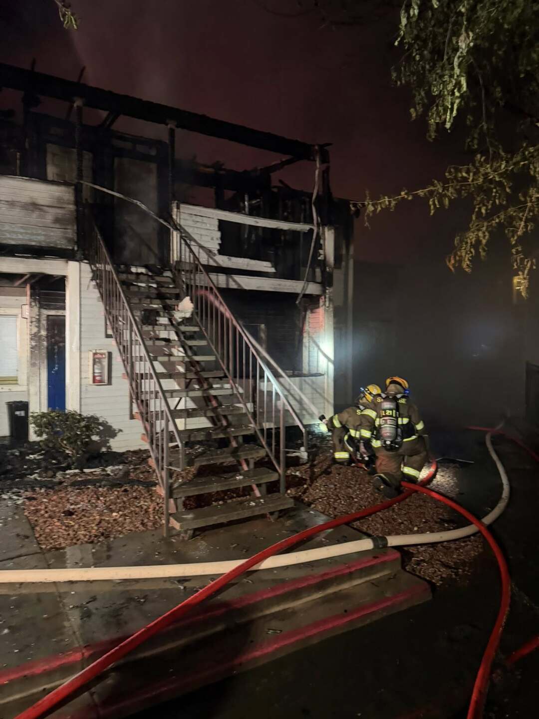 An image from the scene released by St. George Fire Department. It is dark. The apartment building is now a hollow, charred frame. Smoke billows out from within as firefighters continue spraying with the hose.