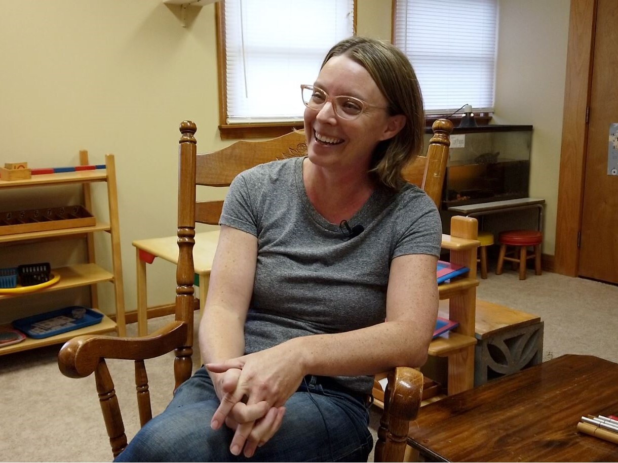 Katie McGoron sits in a rocking chair in the preschool classroom at Shine Nurture Center. She is smiling and had shoulder length, dark blonde hair and glasses.