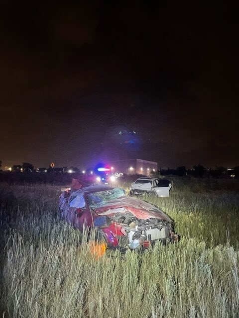 An image of the scene released by Utah Highway Patrol. It's night. The damaged Mustang sits in a patch of vegetation, the hood is dented, and the engine components are visible the windshields of the car are cracked and shattered. Emergency vehicle lights shine in the background.