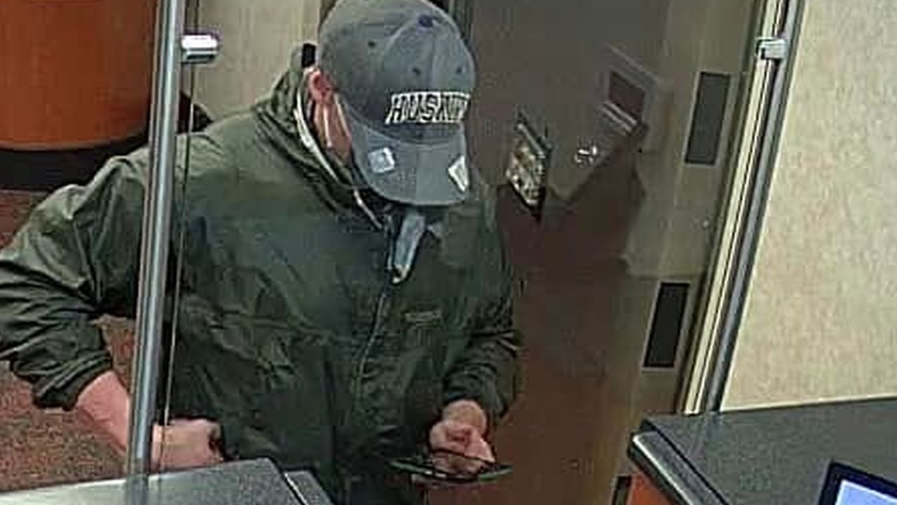 Bank robber wearing blue jeans, green jacket, and baseball cap with “Huskies” on it.