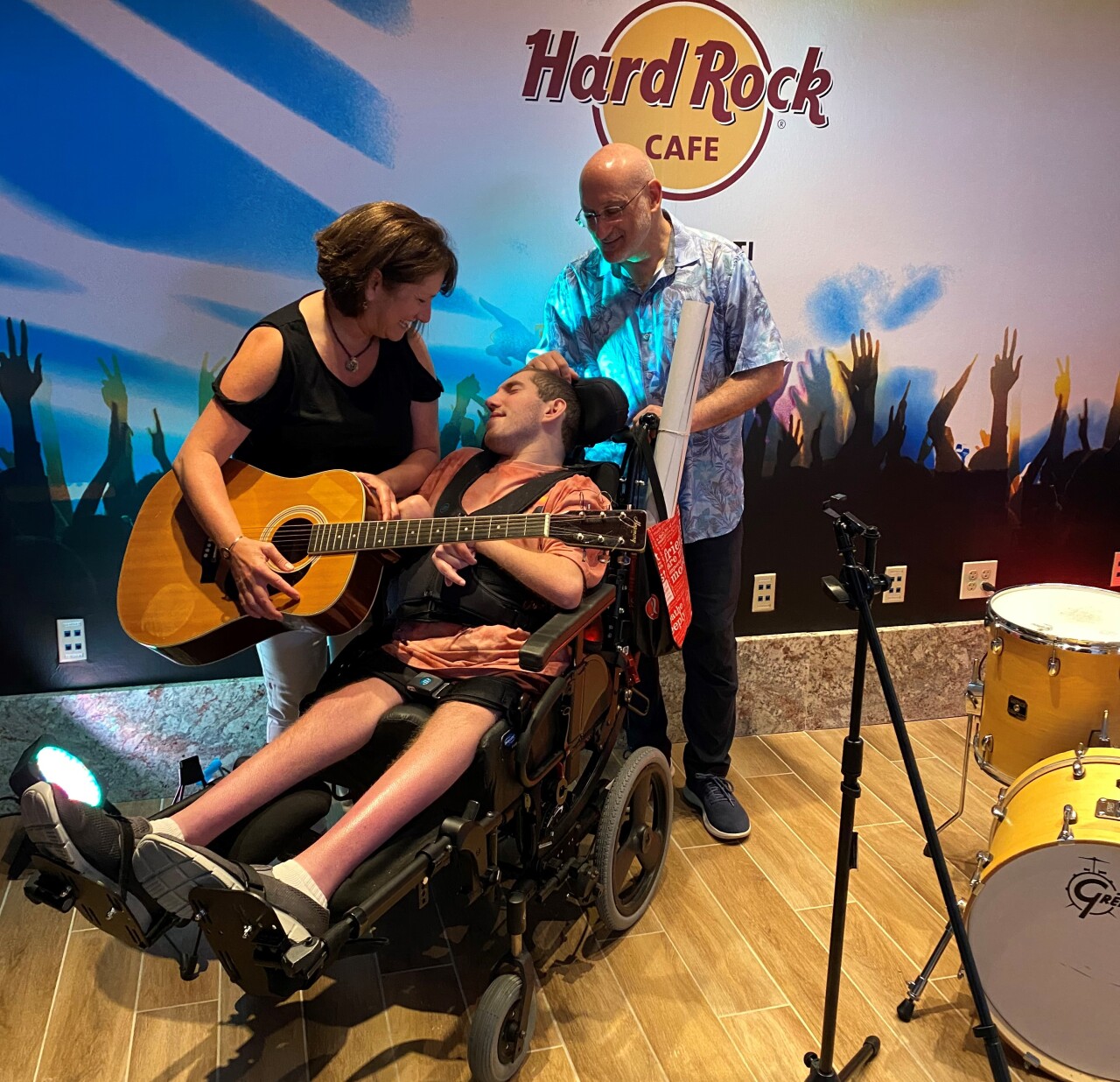 In this photo, Alexia Kadish is holding a guitar near her son, Ethan, who is seated in his wheelchair. Scott Kadish, Ethan's dad, is standing behind Ethan holding the wheelchair.
