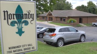 A sign for Normandy, Missouri, is in the foreground with cars and Normandy City Hall in the background.