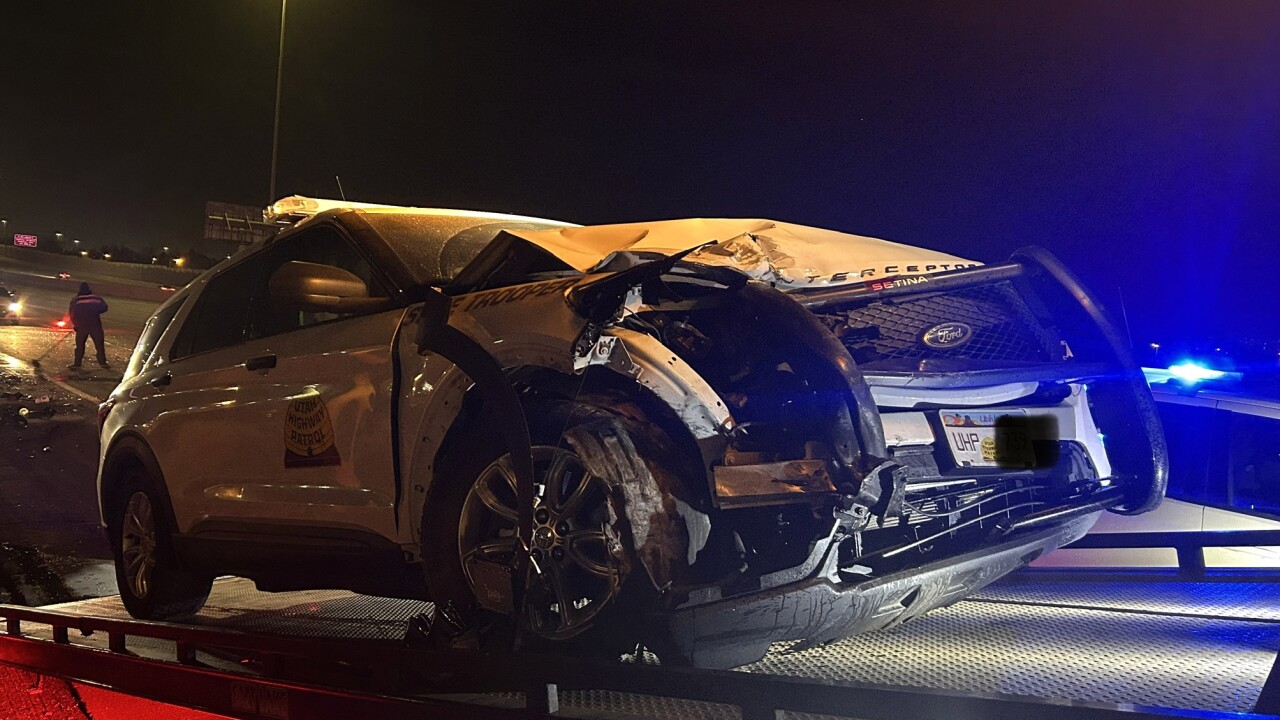 A photo of the scene released by Utah Highway Patrol. It's night. The damaged patrol vehicle is hoisted onto the bed of a tow truck. It shows visible front end damage.