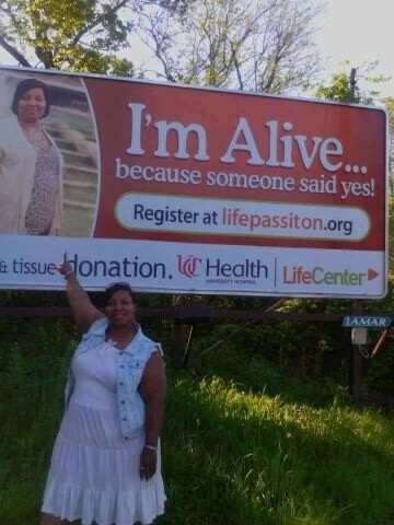 Loretta Davis, wearing a light blue denim vest over a white and gray dress, stands in front of a red billboard displaying her picture and text reading, "I'm alive... because someone said yes!"